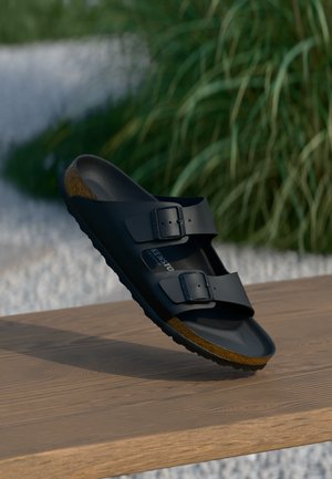Black two-strap sandal with adjustable buckles and cork footbed, suspended above a wooden surface with greenery in the background.