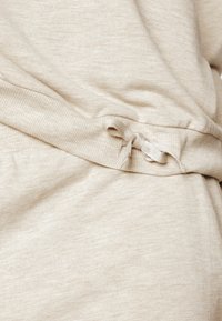 Tissu en coton beige avec une texture douce, présentant un détail de ruban noué à la taille. Le design est simple avec une couleur uniforme.