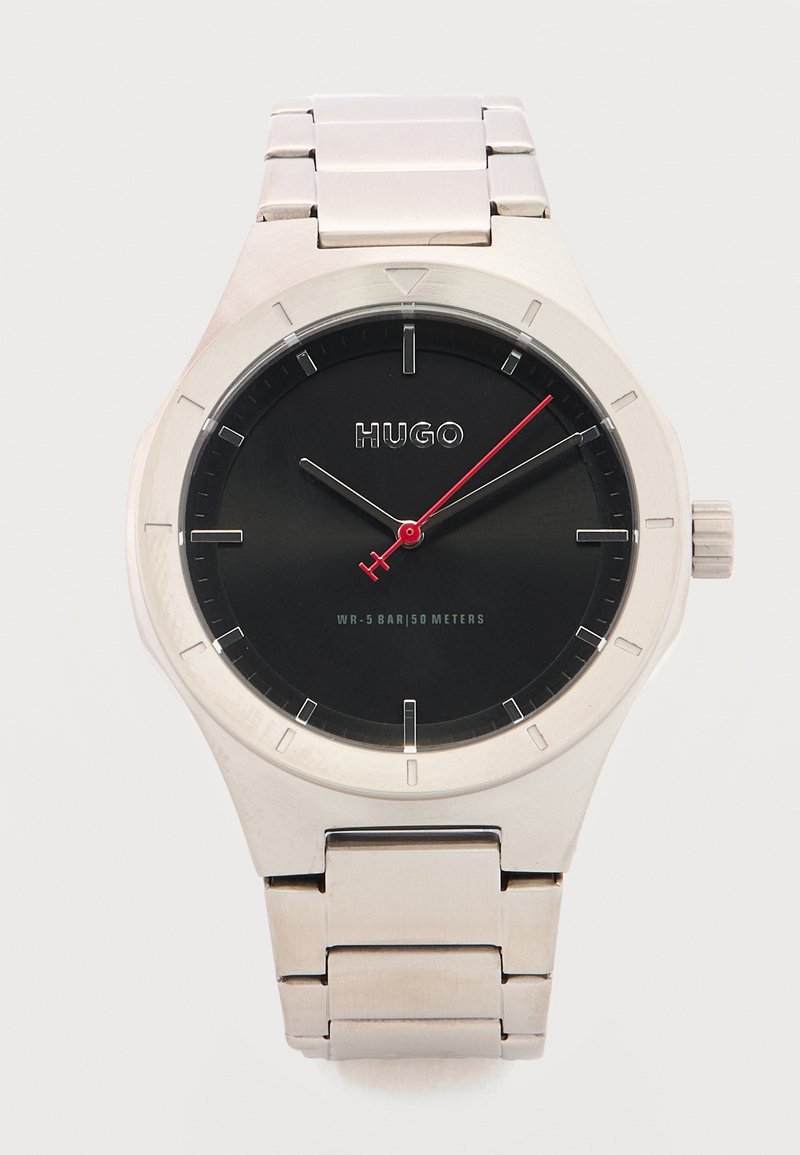 Ασημένιο μεταλλικό ρολόι χειρός με μαύρο καντράν, μινιμαλιστικούς δείκτες ώρας, κόκκινο δείκτη δευτερολέπτ ων και λογότυπο της μάρκας "HUGO" στο καντράν.