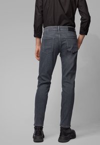 Dark gray denim jeans with a slim fit, featuring two back pockets and a textured waistband. Worn with black sneakers, showing ankle detailing.