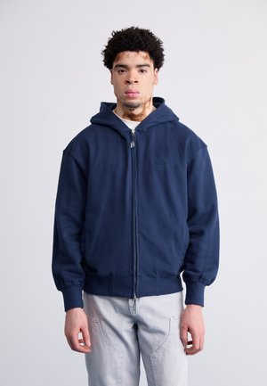 Jūros laivyno zip-up džemperis su traukiama kapuce, priekinėmis kišenėmis ir raištiniais rankogaliais. Ant krūtinės yra mažas logotipas. Minkšta medžiagos tekstūra.