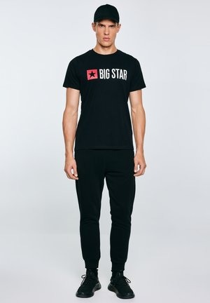 Jauns vīrietis melnā cepurē, melnā "BIG STAR" t-kreklā, melnās sporta biksēs un melnās sporta kurpēs stāv pret vienkāršu gaišu fonu.