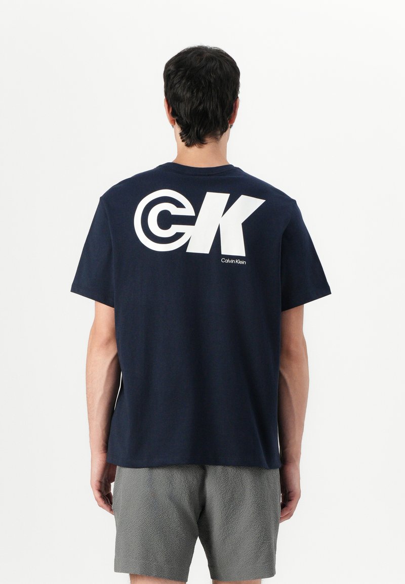 Námornícky modré bavlnené tričko s veľkým bielym logom "CK" na chrbte. Krátke rukávy a voľný strih. Viditeľné sú texturované šedé šortky.