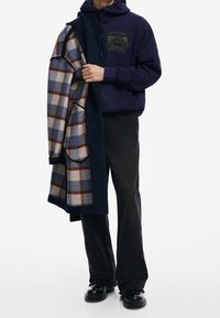 Granatowa bluza z kapturem z grafiką, zestawiona z wełnianym płaszczem w kratę w odcieniach niebieskiego, kremowego i brązowego, noszona z czarnymi spodniami dzwonami i czarnymi botkami.