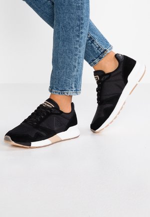 Person trägt schwarze Sportschuhe mit weißen Sohlen und blauen Jeans, steht mit einem leicht angehobenen Fuß.