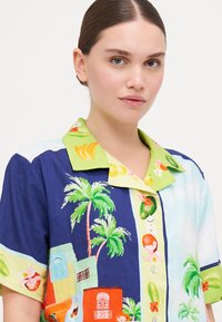 Camicia a maniche corte con una stampa tropicale colorata che presenta palme e frutti. Collo verde, dettaglio con bottoni e vari colori vivaci.
