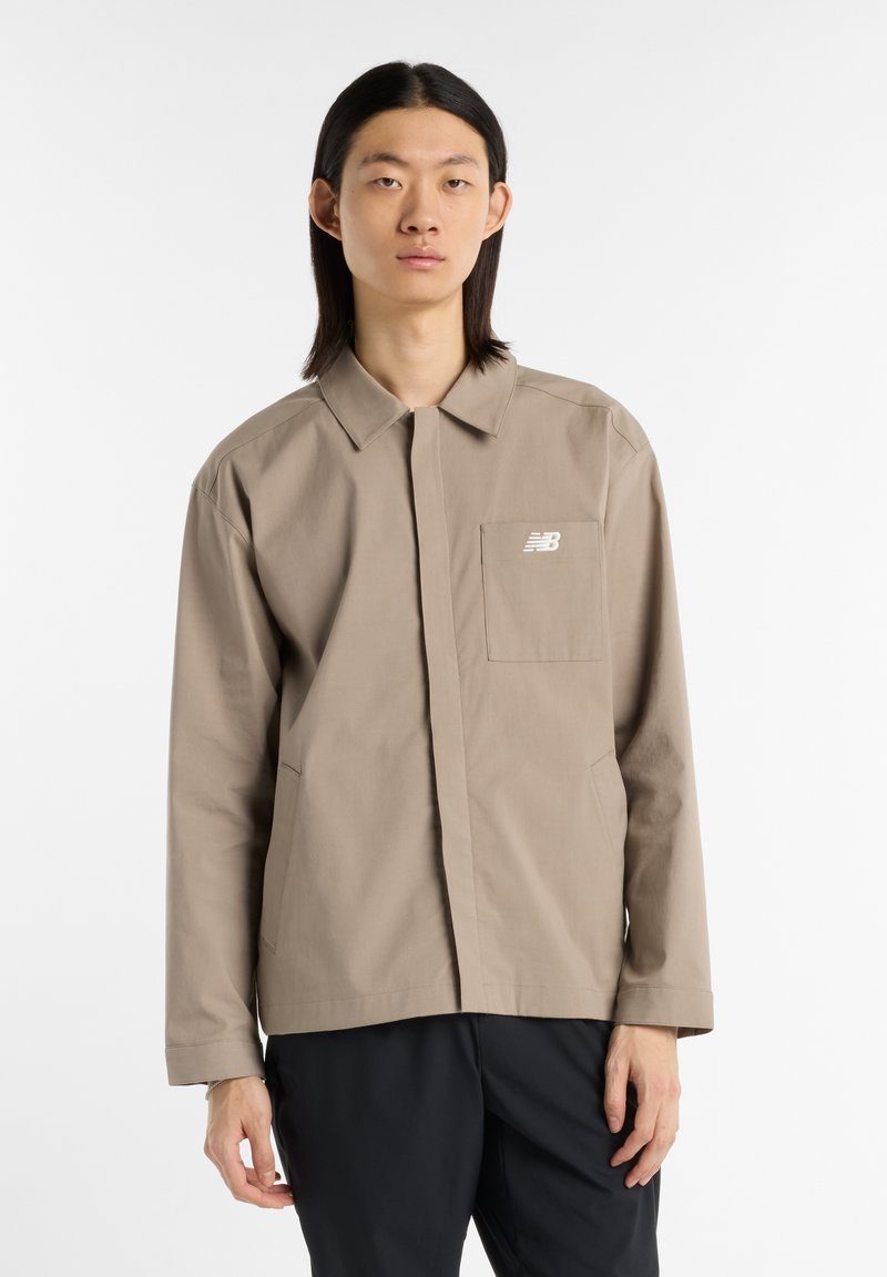 Beige knoopjack met kraag, voorzak en New Balance-logo. Gemaakt van gladde stof, heeft een relaxte pasvorm en lange mouwen.