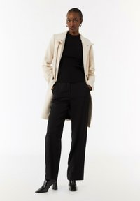 Beiger langer Mantel über einem schwarzen gerippten Pullover, kombiniert mit schwarzen maßgeschneiderten Hosen und schwarzen Absatz-Stiefeletten. Das Model steht mit den Händen in den Taschen.