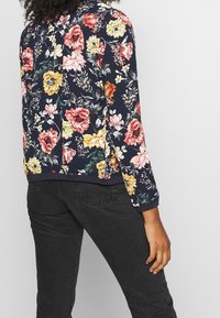 Veste marine à motif floral avec des manches longues, ornée de fleurs roses, jaunes et rouges, et de poignets côtelés. Portée avec un jean foncé.