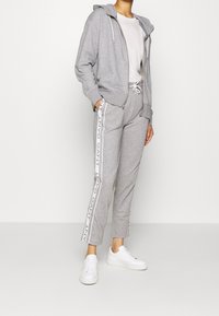 Felpa con cappuccio grigio chiaro con cerniera frontale, dettagli del logo bianchi sulle maniche, abbinata a pantaloni jogger coordinati e sneakers bianche. Tessuto dalla consistenza liscia.