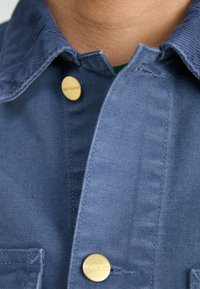 Blått stoffskjorte med teksturert krage, gullknapplukking og synlige sømdetaljer, som fremhever et enkelt, funksjonelt design.