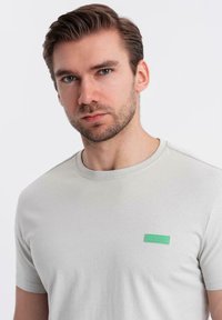 T-shirt in cotone grigio chiaro con maniche corte, dotata di un patch con logo "ombre" verde sul petto e colletto a girocollo.