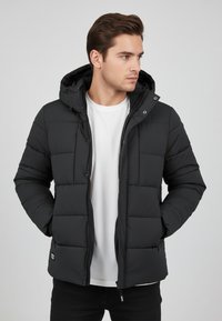 Black puffer jacket with a hood, quilted design, front zipper, two side pockets, and a label on the lower hem. Worn over a white shirt.