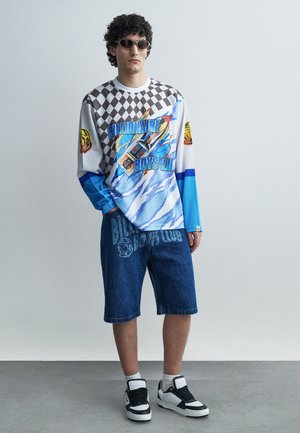 Muž s okuliarmi na slnko, v tričku s dlhým rukávom s nápisom "Billionaire Boys Club", v riflových kraťasoch a čierno-bielych teniskách stojaci pri sivej stene.