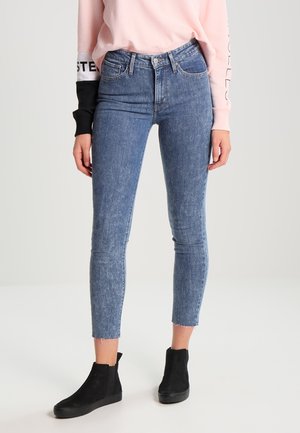 Personne portant un jean skinny bleu taille haute, un sweat-shirt rose clair et des bottines noires, debout sur un fond clair uni.