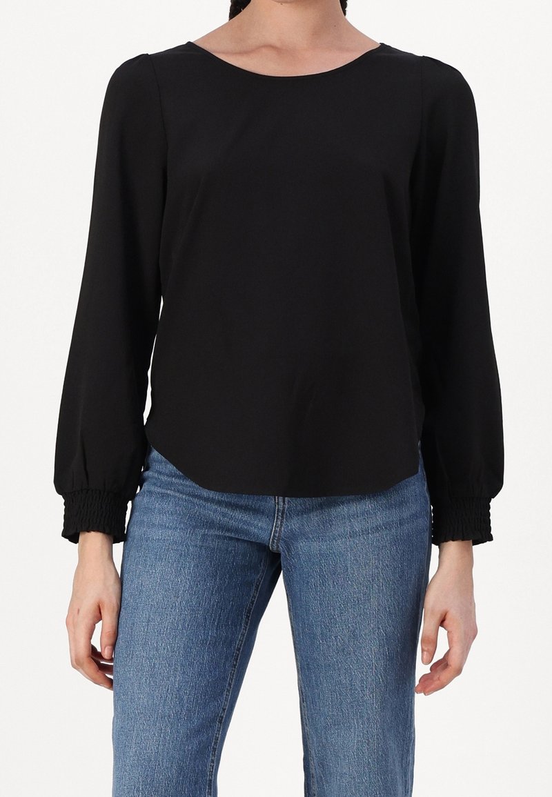 Femme portant un chemisier noir à manches longues avec poignets smockés et un jean bleu, debout devant un fond blanc uni.