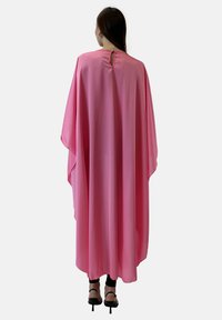 Różowy, zwiewny kaftan o miękkiej fakturze, z detalem w postaci wycięcia w kształcie klucza na plecach i szerokimi rękawami, noszony na czarnych spodniach i na wysokich obcasach.