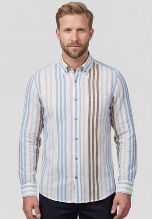 Man met kort blond haar en baard, gekleed in een lang mouwhemd met knopen en verticale blauwe en bruine strepen, staand tegen een effen achtergrond.