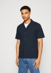 Camisa de manga corta en color marino, hecha de una tela texturizada, con un cuello estilo camp, cierre de botones y dos bolsillos en el pecho. Combina con jeans azules.