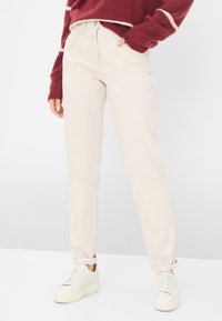 Beige gerade geschnittene Hose mit sichtbaren Taschen, weichem Stoff und subtiler Textur, kombiniert mit weißen Sneakern.