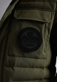 Detailní záběr na kapsu olivově zelené bundy s černou kulatou vyšitou nášivkou zobrazující hory a stromy, označenou "Jacket Summit Division."