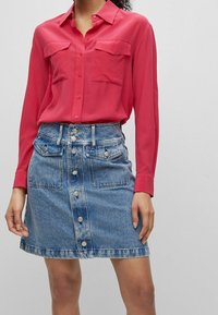 Helles pinkes Hemd mit Knopfleiste und zwei Brusttaschen, kombiniert mit einem hellblauen Jeansrock mit Knopfverschluss an der Vorderseite und zwei Seitentaschen.