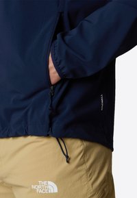 Marinfärgad vindjacka med elastiska muddar, sidofickor med dragkedja och justerbar dragsko i nederkant; tillsammans med beige shorts med en vit logotyp.