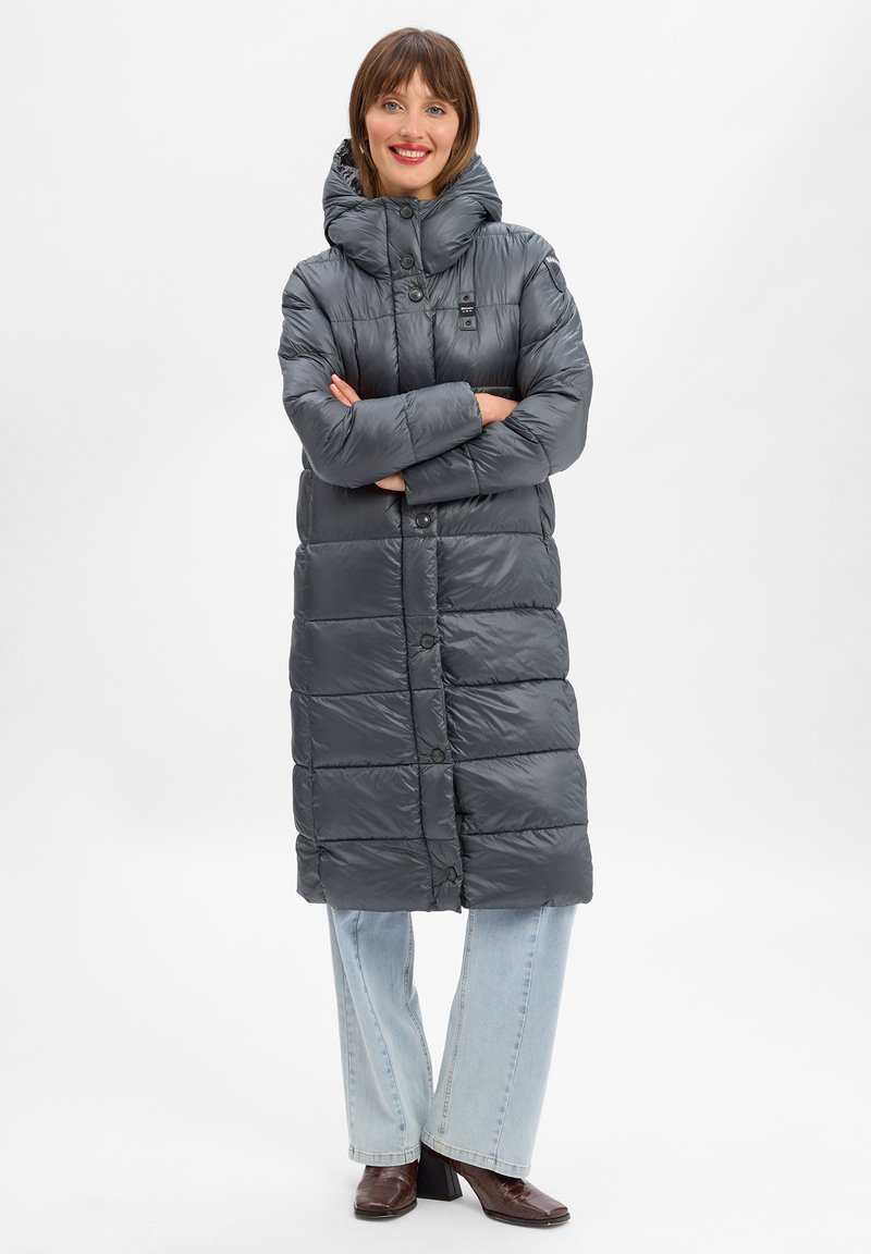 Manteau matelassé gris avec capuche, doté d'une fermeture à boutons et d'un design matelassé. Porté avec un jean bleu clair et des bottes à talons marron.