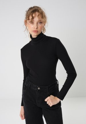 Crna majica s turtleneck izrezom i dugim rukavima, rebraste teksture, uskog kroja; nosi se uz crne traperice visokog struka, koje imaju detalj s gumbom.