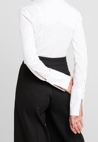 Person in schwarzen hochtaillierten Hosen und einem weißen langärmligen Shirt mit verlängerten Manschetten, die die Hände hinter dem Rücken verschränkt.