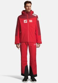 Rote wasserdichte Jacke mit Kapuze und passenden Hosen. Auffällige schwarze Akzente an den Hosenbündchen. Das Jacket verfügt über Logos. Das Material scheint strapazierfähig zu sein.