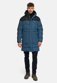 Gepolsterter Wintermantel mit einem dunkelblauen Körper, schwarzen Schultern, Kapuze mit Kordelzug, zwei Fronttaschen und strukturiertem Stoff. Getragen über schwarzen Hosen und Stiefeln.