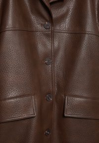 Sezione della giacca in pelle marrone con texture, dotata di quattro bottoni scuri e due tasche rettangolari con patta visibili.