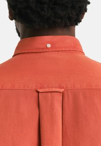 Kurzarmpullover aus Korallengewebe mit glatter Textur. Verfügt über einen knöpfbaren Kragen und einen Faltenwurf von der Schulter, der eine lässige Passform bietet.