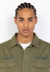 Olive green long-sleeve shirt with button closure, two chest pockets, and embroidered patches reading "US 740N" and "SCHOTT NYC."