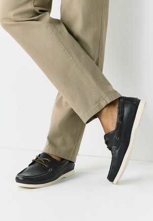 Osoba v béžových kalhotách a černých kožených lodních botách s bílými podrážkami stojí na bílém podlaze s jednou nohou překříženou přes druhou.