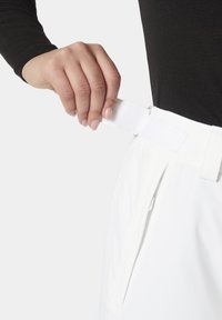 Bílé šortky s nastavitelným pasem na suchý zip, se bočními kapsami a hladkou texturou tkaniny. Zobrazeno ruční nastavení pasu.