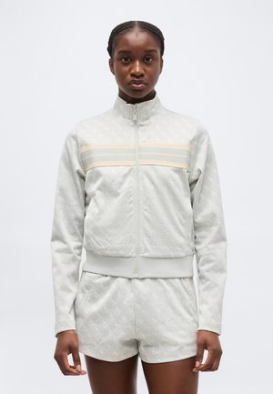 Woman facing forward wearing light gray patterned zip-up jacket with beige stripe and matching shorts on plain background.