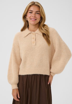 Jeune femme aux cheveux blonds porte un pull en maille beige avec de gros boutons et des manches bouffantes, associé à une jupe marron, souriante à la caméra.