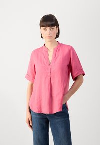 Camicetta rosa in lino a maniche corte con colletto alla cinese, vestibilità morbida e leggeri spacchi laterali. Indossata con jeans blu in denim.