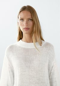 Maglione bianco lavorato a maglia con scollo rotondo e vestibilità ampia. Presenta una texture a trama aperta, maniche lunghe e dettagli sottili al collo.