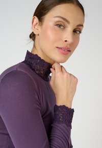 Maglietta viola a maniche lunghe con colletto in pizzo strutturato e polsini in pizzo coordinati. Il tessuto è liscio con una silhouette aderente.