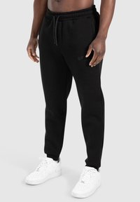 Černé tepláky vyrobené z měkké tkaniny, s elastickým pasem na šňůrku, zúženými nohavicemi a diskrétním logem na boku.