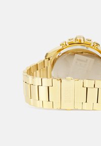Orologio in acciaio inossidabile dorato con bracciale a maglie, cassa rotonda, pulsanti doppi sul lato e fondello trasparente. Logo inciso sulla chiusura.