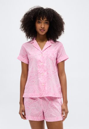 Woman with curly hair wearing pink paisley button-up short-sleeve shirt and matching shorts, standing against a plain light background.