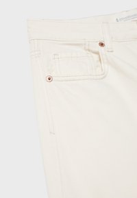 Gros plan sur un jean denim écru montrant la poche avant, la poche monnaie, la boucle de ceinture et les rivets en cuivre sur fond clair.