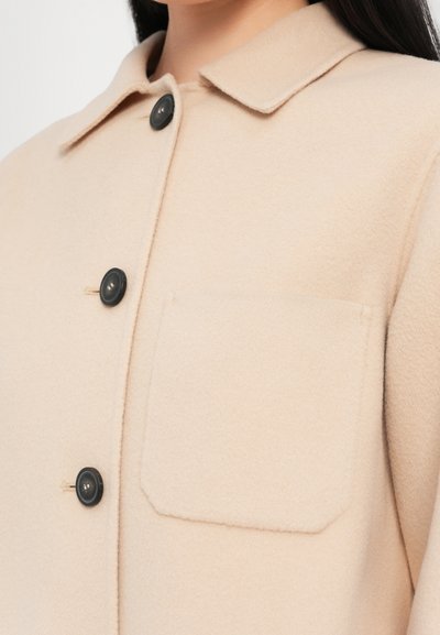 Chaqueta de mezcla de lana beige con collar plano, botones negros y un bolsillo en el pecho. Textura suave, diseño ligeramente oversized.