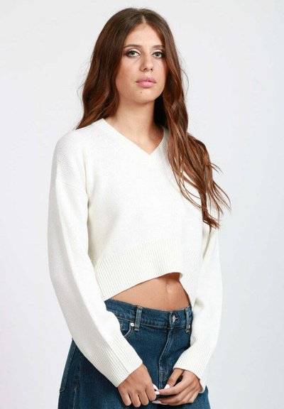 Giovane donna con lunghi capelli castani che indossa un maglione corto bianco con scollo a V e jeans blu a vita alta su uno sfondo semplice.