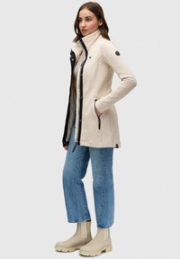Béžová vonkajšia bunda s čiernymi akcentmi, zipsovými vreckami, vysokým golierom a priliehavými rukávmi, nosená s modrými džínami a béžovými členkovými topánkami.
