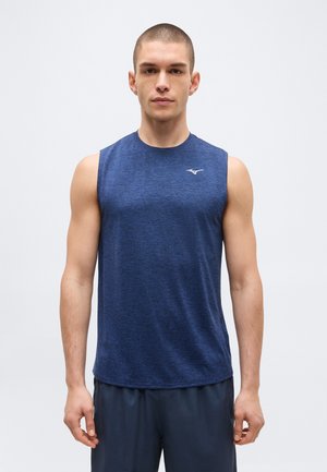 Młody mężczyzna z krótkimi włosami, ubrany w niebieską sportową koszulkę bez rękawów i ciemne szorty, stoi na jasnym, jednolitym tle.